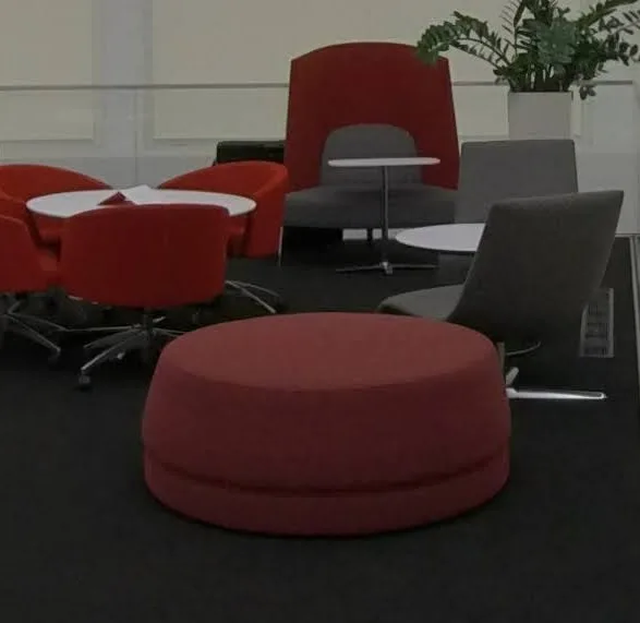 Grand pouf rond recouvert de tissu rouge, d’environ un mètre de diamètre, posé sur le tapis foncé du dôme parmi des fauteuils rouges et gris